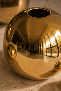 A polished metallic gold decorative sphere sculpture with a circular opening at the top, shown in a close-up studio-style shot on a stone-like surface.