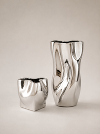 Two modern metallic silver ceramic vases with organic, wavy sculptural shapes, displayed on a neutral warm beige background.