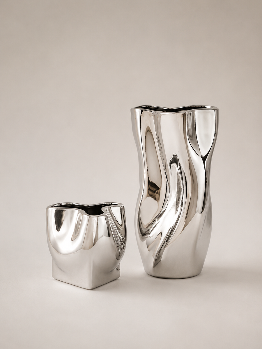 Two modern metallic silver ceramic vases with organic, wavy sculptural shapes, displayed on a neutral warm beige background.