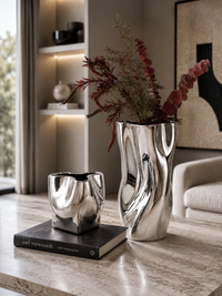 Two modern metallic silver vases with organic wavy contours, one larger holding dried red foliage and one smaller empty vessel, displayed on a stone tabletop in a styled interior setting.