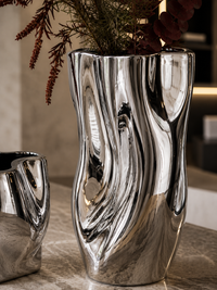 A tall, wavy metallic silver ceramic vase with a reflective, organic surface, holding dried brown and green plant stems, set on a marble countertop.