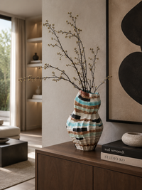 A ribbed ceramic vase with a wavy pattern of turquoise, brown, black, and cream, holding bare branches, styled on a dark wood console table in a modern living room.