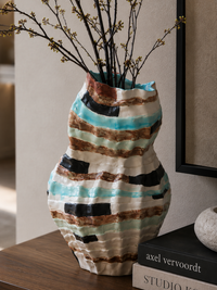 A handmade ceramic vase with a textured, hand-painted pattern of turquoise, brown, black, and cream stripes, holding dried branches, styled on a wooden table next to books.