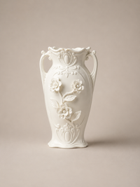 A white ceramic vase with ornate relief floral details and two side handles, centered against a warm beige studio background.