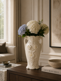 An ornate cream-colored ceramic vase with embossed floral details and side handles, filled with white and blue hydrangeas, displayed on a dark wood console table in a styled living room.