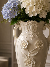 An ornate cream-colored ceramic vase with embossed floral details and two handles, filled with white and blue hydrangeas and green foliage.