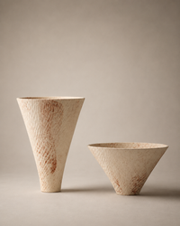 Two handmade ceramic vessels with a textured, speckled beige finish and subtle reddish-brown speckles, displayed on a neutral warm gray background.