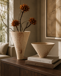 A textured beige ceramic vase with dried orange-brown flower stems and a matching shallow bowl, styled on a dark wooden cabinet in a warm, sunlit room.