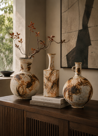 Three decorative ceramic vases with a distressed marbled finish in beige, brown, and cream tones, styled on a wooden console table in a warm interior setting.