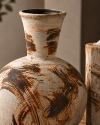 A close-up of a round ceramic vase with a flared neck, featuring a textured off-white glaze with dark brown and rust-colored brushstroke patterns.