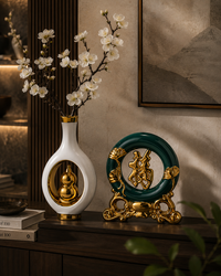 A decorative white vase with a gold-toned interior and gold base, holding white blossoms, displayed beside a green and gold ornamental sculpture on a dark wooden console in an elegant interior setting.