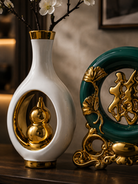 A white ceramic vase with gold trim and a gold gourd motif inside, styled on a wooden surface next to a green and gold decorative plaque in an indoor setting.