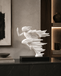 A white sculptural figurine of a stylized dancer or winged form displayed on a black rectangular base, set on a dark marble cabinet in a modern interior with framed wall art and shelving.