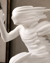 A white sculptural figurine of a winged female figure with flowing hair and draped fabric details, shown in a close-up studio-style shot against a soft neutral background.
