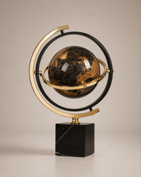 A decorative globe sculpture with a black and gold marbled sphere, surrounded by a gold and black ring frame, mounted on a polished black marble base against a neutral beige background.