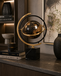 A decorative tabletop globe with a black and gold marbled sphere, gold-toned ring arms, and a black marble base, styled on a dark marble shelf in an interior setting with books and a framed print.