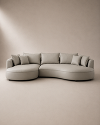 A modern curved sectional sofa upholstered in light taupe fabric with matching throw pillows, set against a minimalist beige studio background.