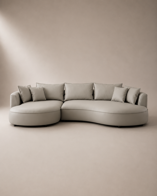 A modern curved sectional sofa upholstered in light taupe fabric with matching throw pillows, set against a minimalist beige studio background.