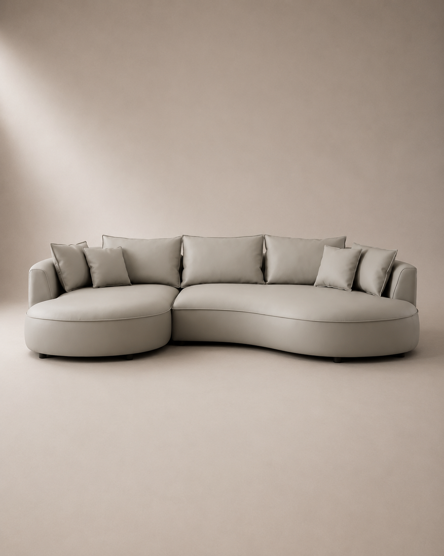A modern curved sectional sofa upholstered in light taupe fabric with matching throw pillows, set against a minimalist beige studio background.