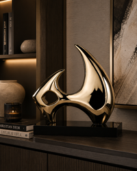 A polished gold abstract metal sculpture on a black rectangular base, displayed on a dark wood console table in a modern interior setting.
