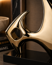 A polished gold abstract metal sculpture on a black base, photographed in a styled interior setting with books and a bowl in the background.