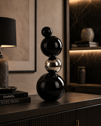A modern black and silver stacked sphere sculpture displayed on a dark wood console table in a sophisticated, dimly lit interior setting.