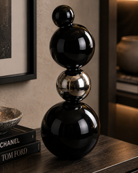 A glossy black and metallic silver stacked sphere sculpture displayed on a dark wooden table next to books in a modern, warmly lit interior setting.