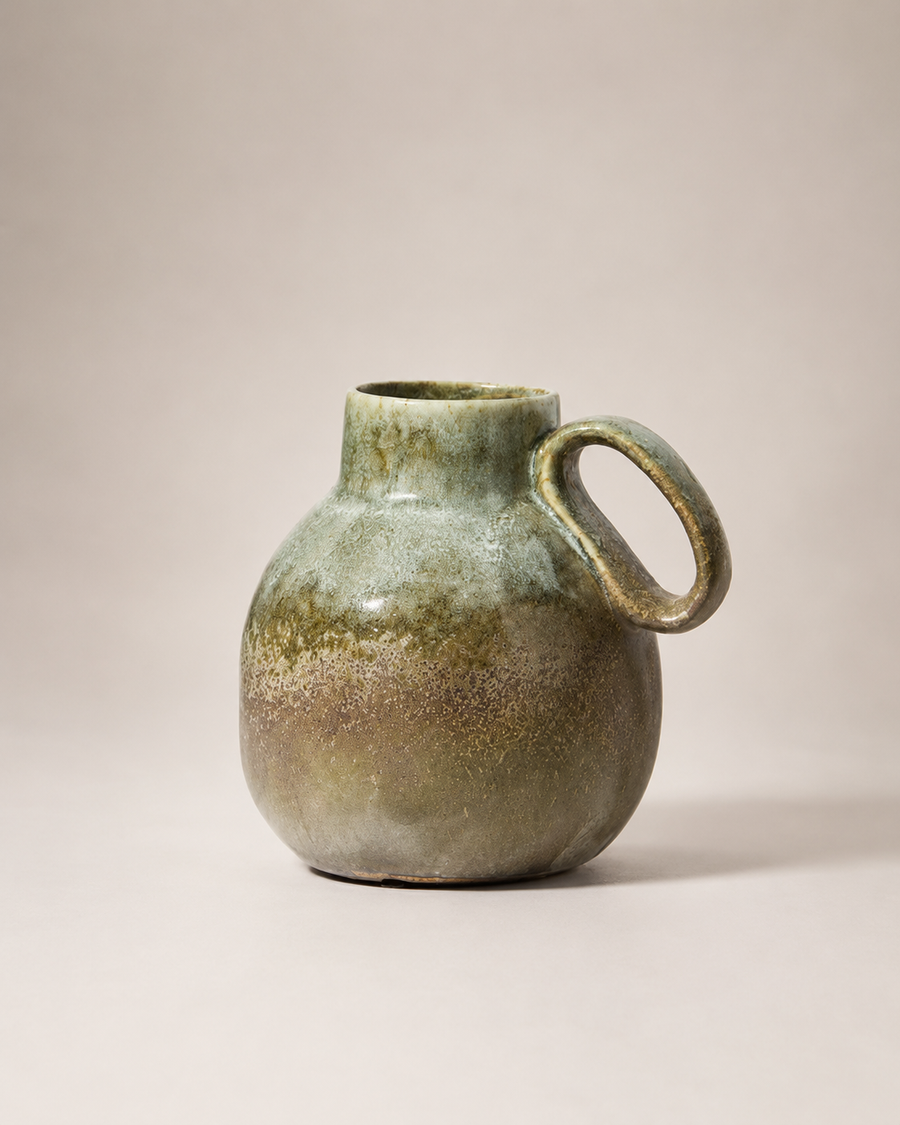 A handmade ceramic pitcher with a rounded body, a small loop handle, and a short neck, featuring a textured glaze that transitions from olive green at the top to a warm brown at the base, set against a solid light beige background.