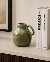 A small glazed ceramic pitcher with a loop handle sits on a light stone surface beside two upright books against a warm beige wall.