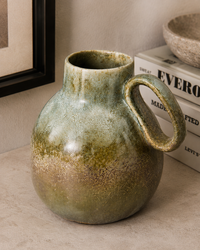 A ceramic pitcher vase with a rounded body and loop handle, featuring a textured glaze that transitions from olive green at the top to a warm brown at the base, styled on a tabletop next to books.
