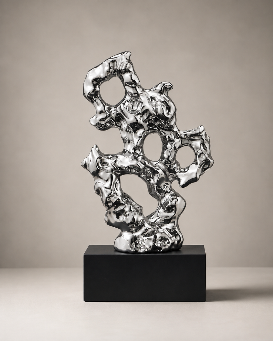 A polished silver abstract sculpture with organic, branching forms displayed on a matte black rectangular pedestal against a warm beige studio background.