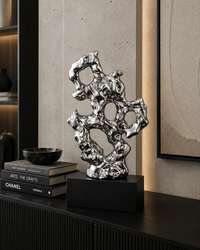 A polished silver abstract metal sculpture with organic, flowing contours displayed on a black rectangular pedestal atop a dark console table. The setting includes a textured concrete wall, a stack of books, a small black bowl, and a framed artwork in the background.
