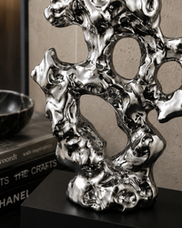 A polished silver abstract sculpture with organic, fluid contours and hollow circular openings, displayed on a black pedestal in an indoor setting with books and a bowl in the background.