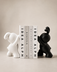 Two stylized bear-shaped bookends, one matte white and one matte black, holding a row of four books including 'IDEA ALPH' and 'KINFOLK' against a warm beige background.