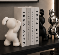 A styled interior shelf scene with a white ceramic bear figurine, a black book titled 'The Kinfolk Entertainer', a black bear figurine, and a shiny silver balloon-boy style rabbit figurine on a dark wooden shelf.