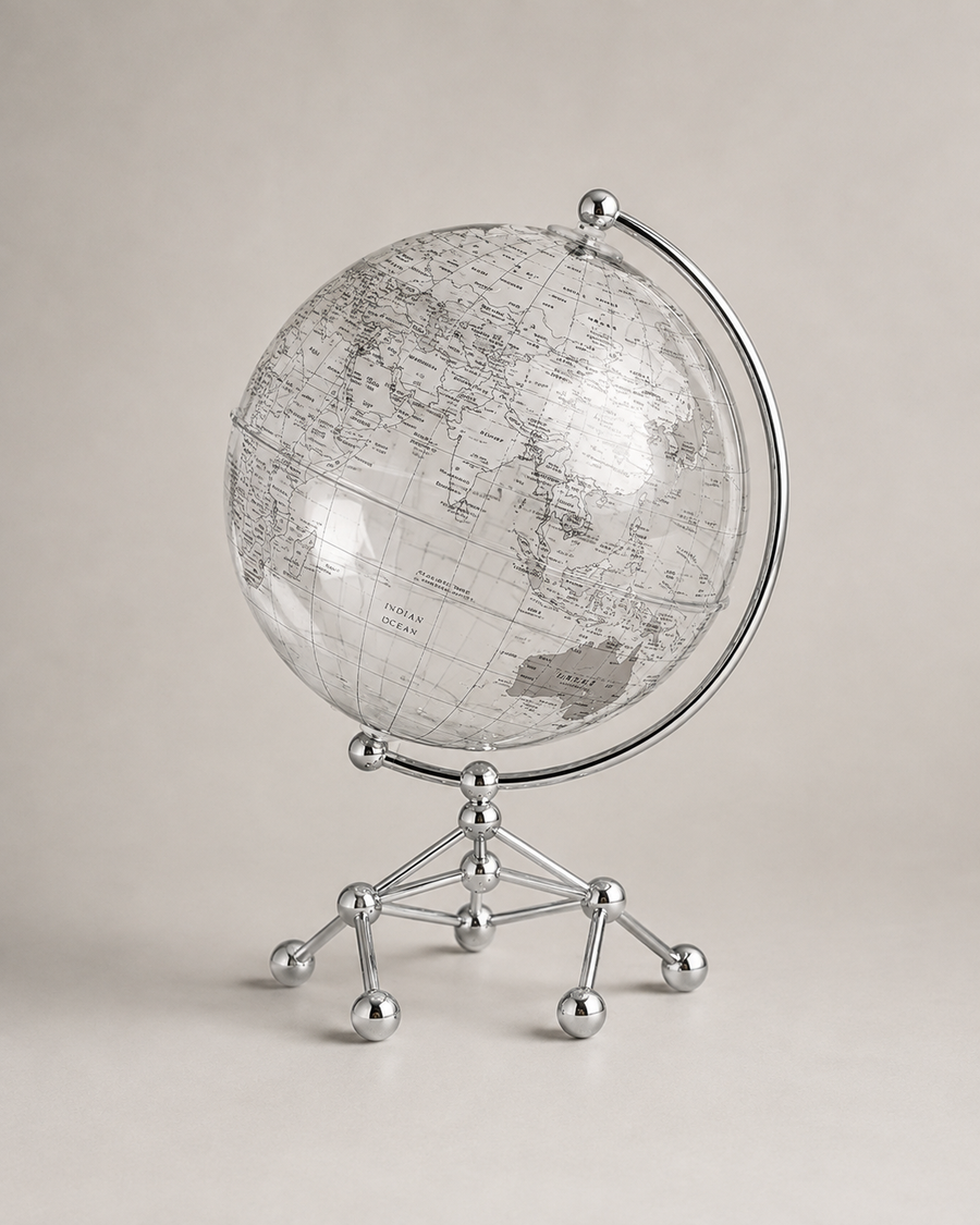 A clear glass globe with a vintage-style gray map print, mounted on a polished silver metal stand with a curved arm and five legs, set against a neutral beige background.