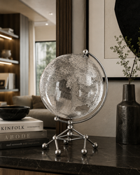 A clear glass globe with a silver metal stand and ring, displayed on a dark marble tabletop in a modern, upscale living room setting.