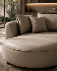 A close-up of a modern round daybed or chaise lounge in taupe leather, styled with three matching square throw pillows in a contemporary living room setting.