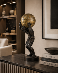 A dark bronze sculpture of a muscular figure carrying a large gold globe on his shoulders, displayed on a dark console table in a modern, stylishly decorated room.