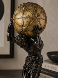 A classical-style bronze sculpture of a muscular figure carrying a large gold globe on his shoulders, displayed indoors against a neutral wall with framed art and books in the background.