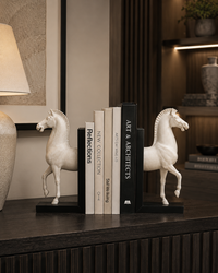 A pair of white horse bookends holding a set of books on a dark wooden console table in a modern, warmly lit interior setting.