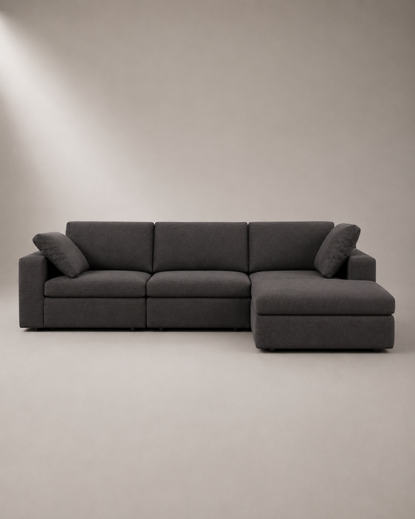 A modern three-seat sectional sofa with a right-side chaise lounge, upholstered in dark gray fabric, set against a minimalist beige studio background.