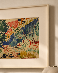 Framed abstract landscape painting with expressive brushstrokes in green, blue, red, orange, and yellow on a cream background, shown hanging on a light wall.
