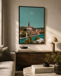 A framed travel poster of Bern, Switzerland, hanging on a beige wall in a styled living room setting with a wooden console table, a lamp, and a sofa.