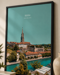 A framed vertical art print of Bern, Switzerland, featuring a scenic cityscape with the cathedral spire and river under a teal sky, styled on a beige wall next to a ceramic vase.