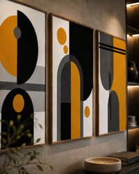 Three framed abstract geometric wall art prints with black, mustard yellow, gray, and white shapes, displayed on a textured gray wall in a modern interior setting.