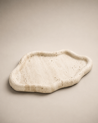 An organic-shaped travertine stone tray with a textured, speckled natural surface, displayed on a neutral beige background.