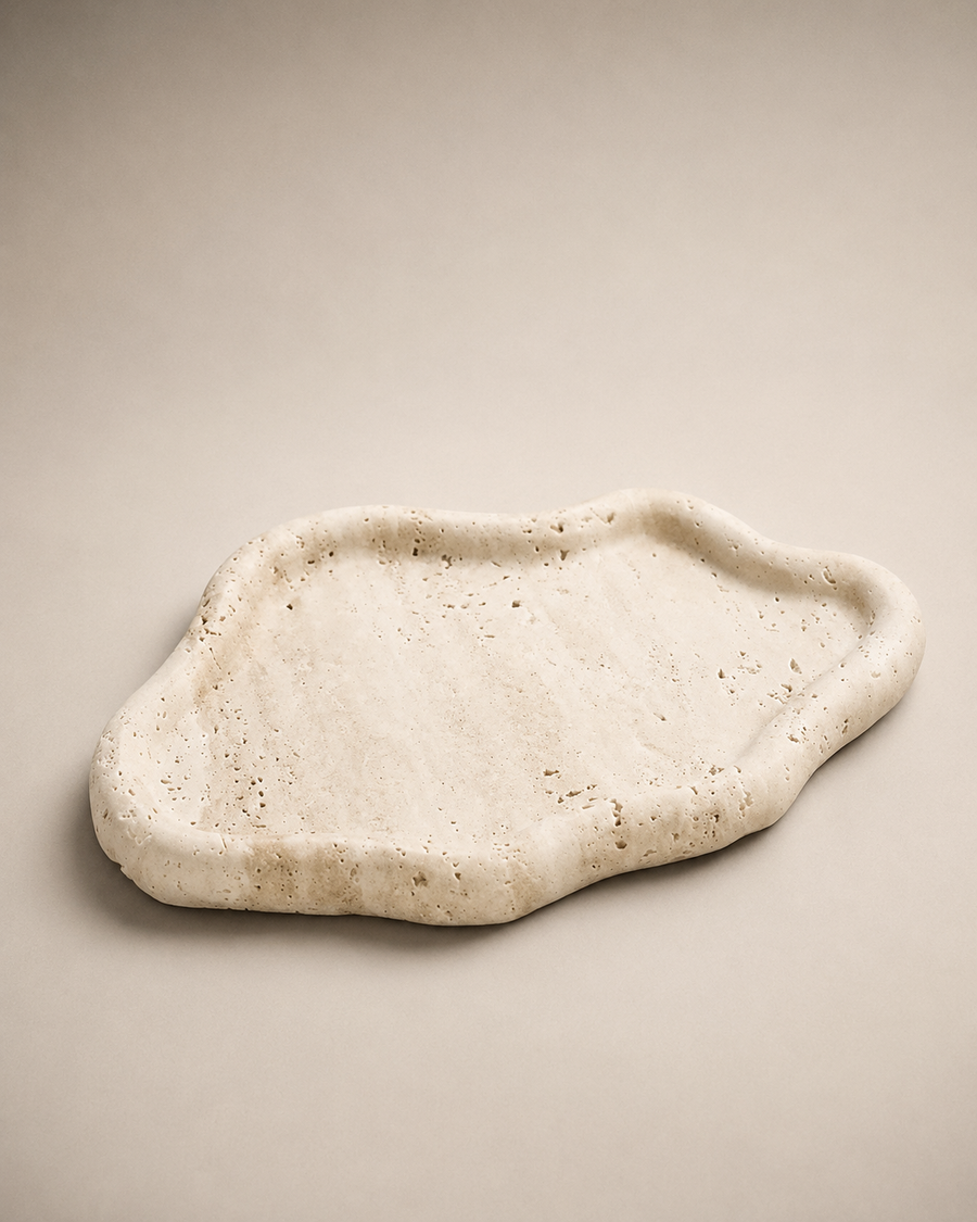 An organic-shaped travertine stone tray with a textured, speckled natural surface, displayed on a neutral beige background.