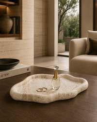 An organic-shaped travertine stone tray with a speckled cream and beige texture sits on a dark wood table in a modern, warm-toned living space. The tray is styled with a small glass perfume bottle and two gold rings.