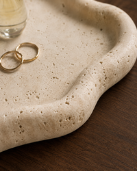 A close-up of a beige travertine stone tray with organic wavy edges, resting on a dark wood surface. Two gold rings and a glass bottle are placed on the tray as styling props.